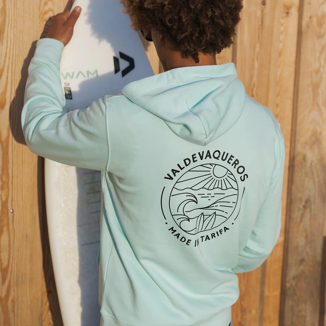 fotografía de Matchu Lopes con la sudadera de Made in Tarifa, con el diseño de Valdevaqueros en un color azul mar y una tabla de kitesurf